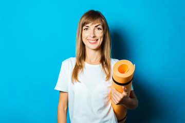 A young woman in a white T-shirt holds a yoga mat on a blue background. The concept of yoga, yoga for beginners, fitness, stretching