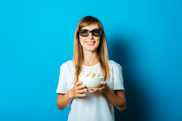 Young beautiful woman in a white T-shirt smiles with glasses for 3D and iMAX holds a plate with popcorn on a blue background. Concept of watching new movies at the cinema or at home
