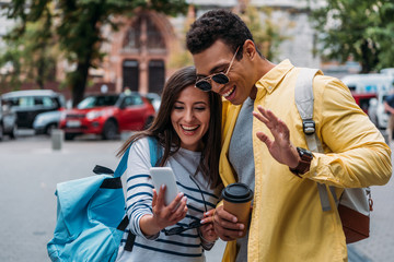 Bi-racial man with paper cup waving hand near woman tacking selfie on smartphone