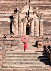 novice buddhist monks with red traditional robes holding red umbrellas walking in a brown buddhist temple in myanmar