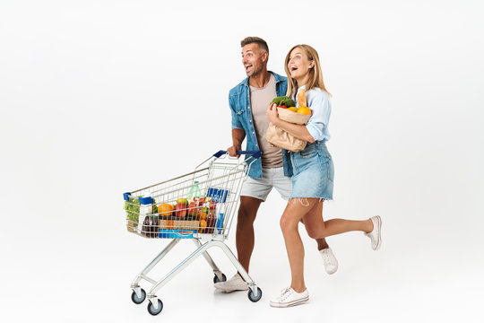Photo of joyful family woman and man holding food products in supermarket and carrying shopping cart isolated over white background