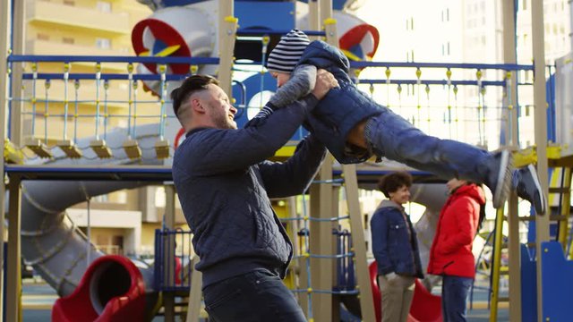 Slowmo Panning Shot Of Cheerful Caucasian Dad Having Fun With Laughing Young Son On Playground, Holding Him, Swinging And Spinning Around Together, And Two Mums Standing And Chatting In Background