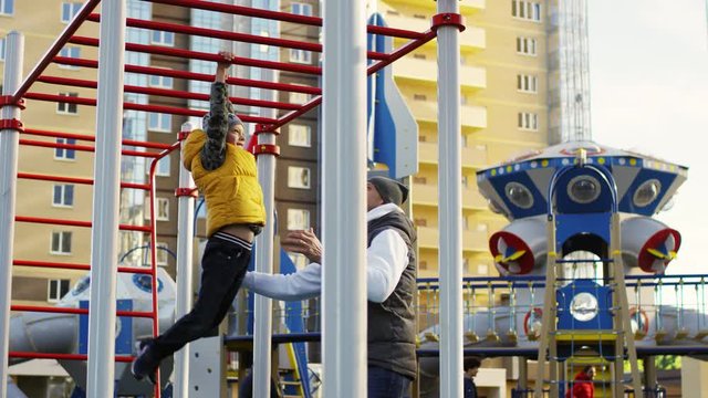 Full shot of fit 6-year-old Caucasian boy exercising on horizontal ladder in playground, swinging on it, gripping crossbars and moving along, while caring dad is standing by and spotting him