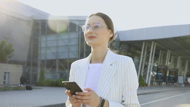 Beautiful young businesswoman wearing white shirt and using modern smart phone while walking at break in the city, professional female employer typing text message on cellphone outside, slow motion