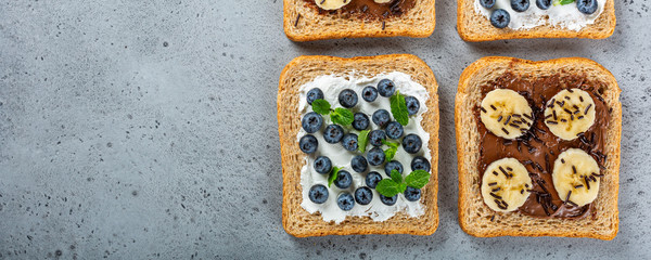 Delicious open sandwiches with goat cheese and blueberries and chocolate and banana on gray table. Breakfast background. Copy space, top view, banner.
