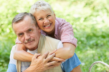 Portrait of happy mature woman embracing her husband while standing behind his back and they smiling at camera outdoors in summer day