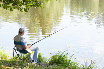 Mature man sitting on chair with fishing rod and fishing near the pond in nature