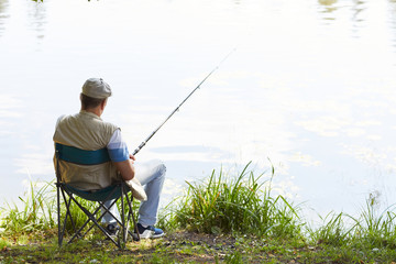 Rear view of mature fisherman sitting on chair with fishing rod and waiting for a catch at the...