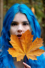 Beautiful young bohemian woman with blue hair happily holding a yellow orange autumn leaf in her...