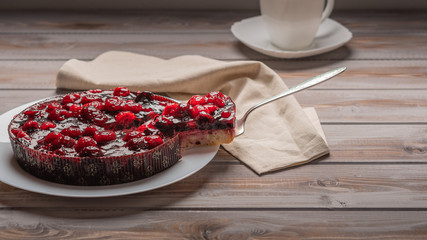 Pie with berries: raspberries, strawberries, currants, on a white plate, next to a piece of cake on the shoulder. On a wooden background, in the background napkin, white cup and saucer