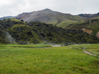 Group of tourist people relaxing in a natural hot spring in thermal baths in Landmannalaugar camp site, Iceland. Grass meadow, lava fields and mountains in background