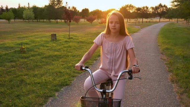 Girl ride bicycle on park alley against the setting sun - slow motion, front view, medium close up
