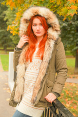 Redheaded woman in coat with fur hood in autumn park with yellow green trees