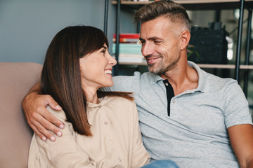 Photo of middle-aged caucasian couple smiling while cuddling on sofa at home