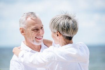 happy senior couple in white shirts embracing and looking at each other under blue sky