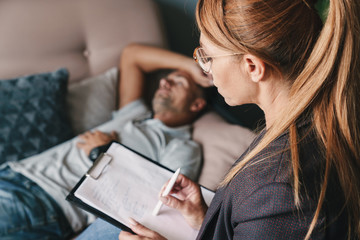 Photo of thoughtful caucasian man lying on sofa and having conversation with psychologist in room