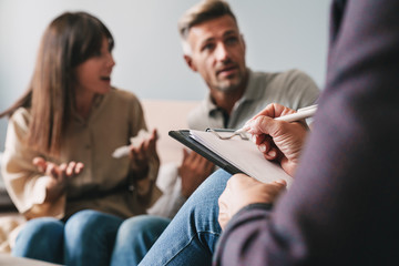 Photo of puzzled irritated couple having conversation with psychologist on therapy session in room