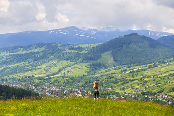 Fototapeta na wymiar Back view of slim young woman standing on grassy valley on background of green mountains on sunny summer day.