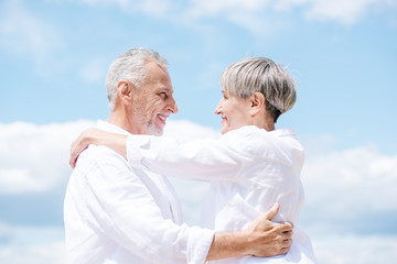 side view of smiling senior couple embracing and looking at each other under blue sky