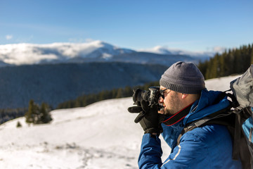 Hiker man tourist photographer in warm clothing with backpack and camera taking picture of snowy valley and woody mountain peaks landscape under blue sky on sunny winter cold day.