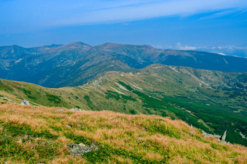 The high mountain range is overgrown with green and yellow grass against the blue sky with clouds