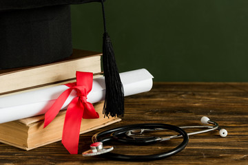 Mortarboard, textbooks, graduation scroll tied with red ribbon and stethoscope 