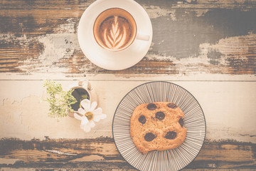 big chocolate chip cookie with a cup of cappunico with latte art on top and a small vase of flowers