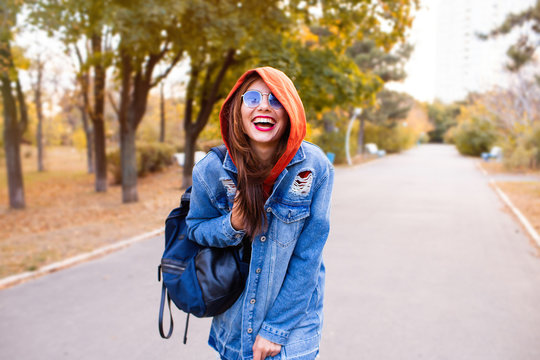 Outdoor fashion portrait of stylish young woman having fun, emotional face , laughing, looking at camera. Urban city street style. Long jeans coat and orange hoodie. Spring or fall outfit.