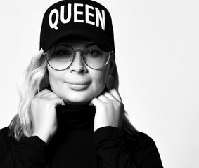Black and white portrait of blonde woman in black cap and big glasses holding her sweater collar with both hands