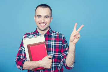 Attractive young school teacher or student with books, man shows sign peace, portrait, blue background, copy space, slogan, toned