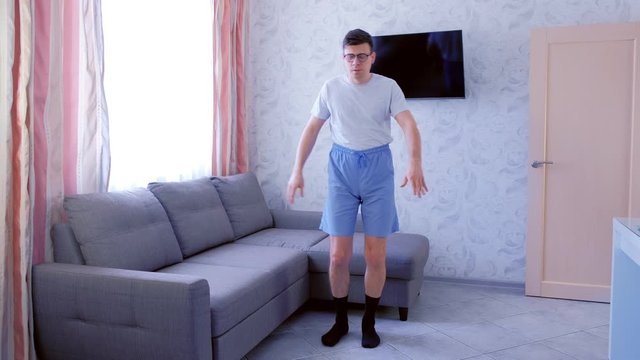 Funny nerd man is making slopes to the legs and touches the legs with his hands, exercise at home in living room. Funny pulls up shorts before exercise. Front view.
