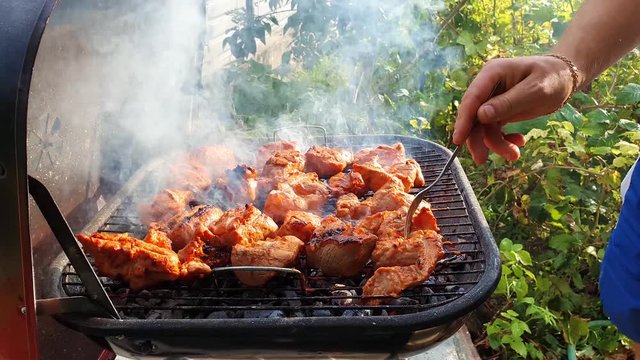 Close up of male hands baking meal outdoors at grill using a fork to turning around meat for not burn out. Barbeque and picnic season concept. Feel the great flavour through the dense steam. 
