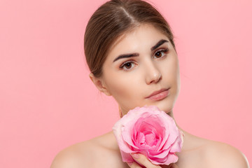 Naklejka premium Close-up portrait of a beautiful young girl looking at the camera holding pink rose flower close to face isolated over pink background.