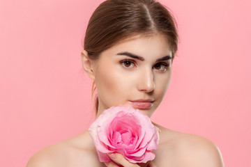 Naklejka premium Close-up portrait of a beautiful young girl looking at the camera holding pink rose flower close to face isolated over pink background.