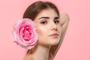 Naklejka premium Close-up portrait of a beautiful young girl looking at the camera holding pink rose flower close to face isolated over pink background.