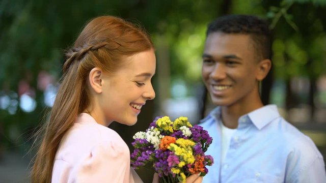 Shy Afro-American boy presenting bouquet of flowers to smiling girl in park