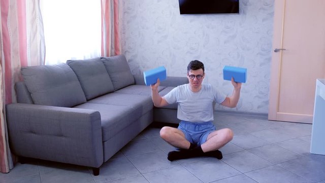 Weak nerd man in glasses is doing exercises for hands with yoga blocks instead of dumbbells sitting on the floor at home. Sport humor concept.