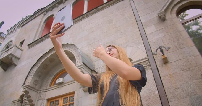 Closeup portrait of young attractive caucasian female with long hair in glasses taking selfies on tablet outdoors on street with wind blowing