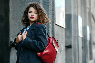 Outdoor waist up portrait of young beautiful curly woman, plus size model with red lips makeup, wearing autumn gray coat, with leather backpack, posing in street of city. Copy, empty space for text