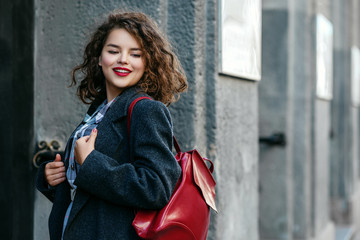 Outdoor waist up portrait of young beautiful happy smiling curly lady, plus size model, wearing autumn gray coat, with leather backpack, posing in street of city. Copy, empty space for text