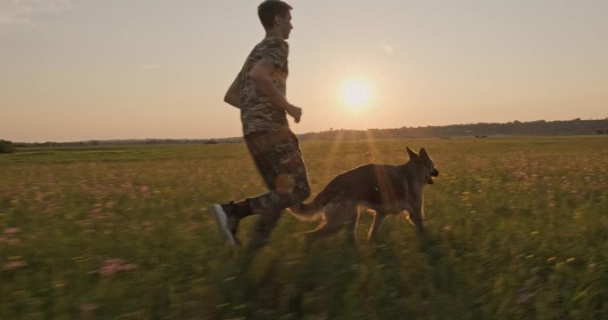 Teenager Is Running With A Dog In Nature While Sunset. 15 Year Old Boy Runs With A Dog Breed German Shepherd. Teen On The Field With Dog. Caucasian Guy Is Playing With His Pet In The Meadow. 