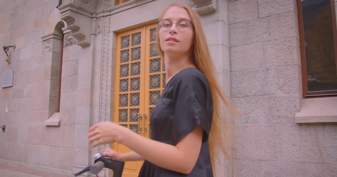 Closeup portrait of young attractive caucasian female with long hair in glasses and black dress holding kick scooter looking at camera outdoors