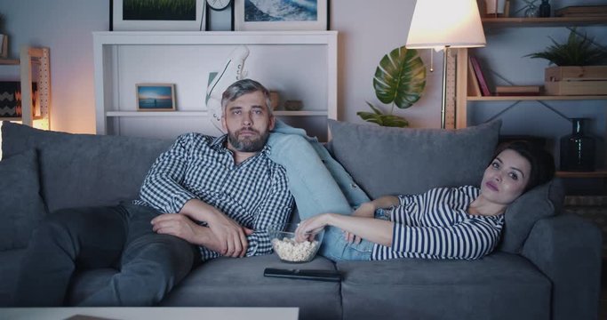 Girl and guy watching TV at night eating popcorn on couch together relaxing in dark room. Entertainment, relaxation and modern people concept.