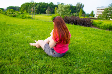 Fototapeta premium Red-haired girl sits with his back on the grass in the park. Vacation and vacation concept.