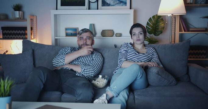 Husband and wife middle-aged people are watching TV at home eating popcorn laughing enjoying modern television at night. Family and leisure concept.