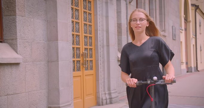 Closeup portrait of young pretty caucasian female with long hair in glasses and dress holding kick scooter looking at camera smiling outdoors