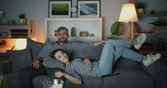 Portrait of girl and guy laughing watching TV at night in cozy apartment sitting and lying on sofa together looking at camera. People, television and fun concept.