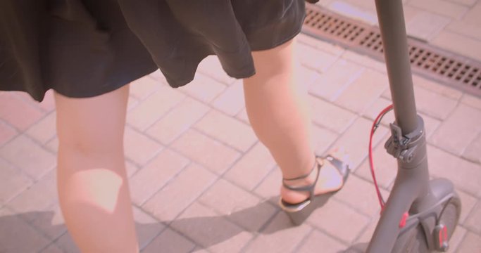 Closeup portrait of young pretty female with long hair in glasses and dress walking with kick scooter happily outdoors