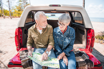 smiling senior couple sitting on car and holding map in sunny day