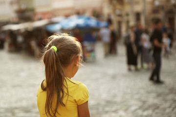 A little girl blonde looks into the distance standing on the street of a medieval European city....